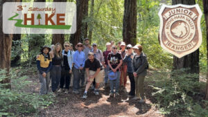 Hikers at the Jr Rangers for Adults hike with the 3rd Saturday Hike logo and a wooden Jr Ranger badge